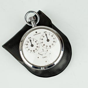 May include: A vintage Junghans stopwatch with a white face, silver casing, and black leather case. The stopwatch features three dials for seconds and minutes, with clear markings. The watch is a classic design, perfect for timing events.