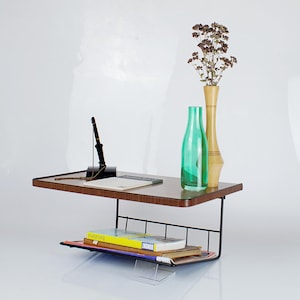 May include: A wall-mounted shelf with a wooden top and a black metal wire rack underneath. The shelf has a green glass bottle and a wooden vase with dried flowers on top. There are books on the rack.