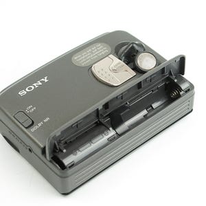 May include: A dark gray Sony portable cassette player with an open battery compartment. The device features a silver latch, a sliding on-off switch, and a Dolby NR label. A small circular sticker with text is positioned near the top edge.