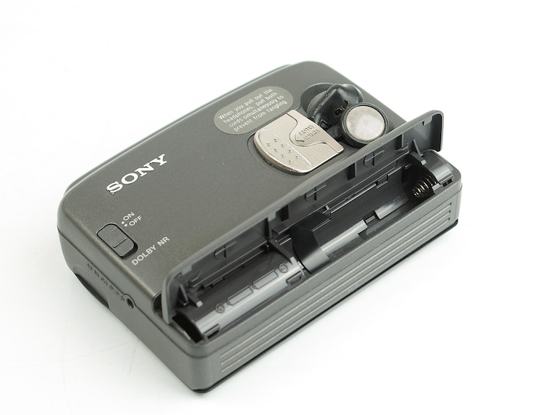 May include: A dark gray Sony portable cassette player with an open battery compartment. The device features a silver latch, a sliding on-off switch, and a Dolby NR label. A small circular sticker with text is positioned near the top edge.