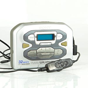 Op de afbeelding: Een zilveren Sony FM/AM Walkman met een zwarte afstandsbediening. De Walkman heeft een blauw LCD-scherm en knoppen zoals "MENU", "REW" en "FF". De tekst "32 PRESETS MEGA BASS" is zichtbaar op het apparaat.
