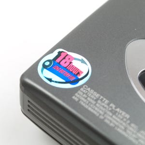May include: A dark gray portable cassette player featuring a circular sticker that reads 18 hours long battery life. The device has a silver-toned accent panel with the text AUTO WIND and AUTO REVERSE printed in blue and black on the casing.