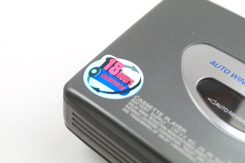 May include: A dark gray portable cassette player featuring a circular sticker that reads 18 hours long battery life. The device has a silver-toned accent panel with the text AUTO WIND and AUTO REVERSE printed in blue and black on the casing.
