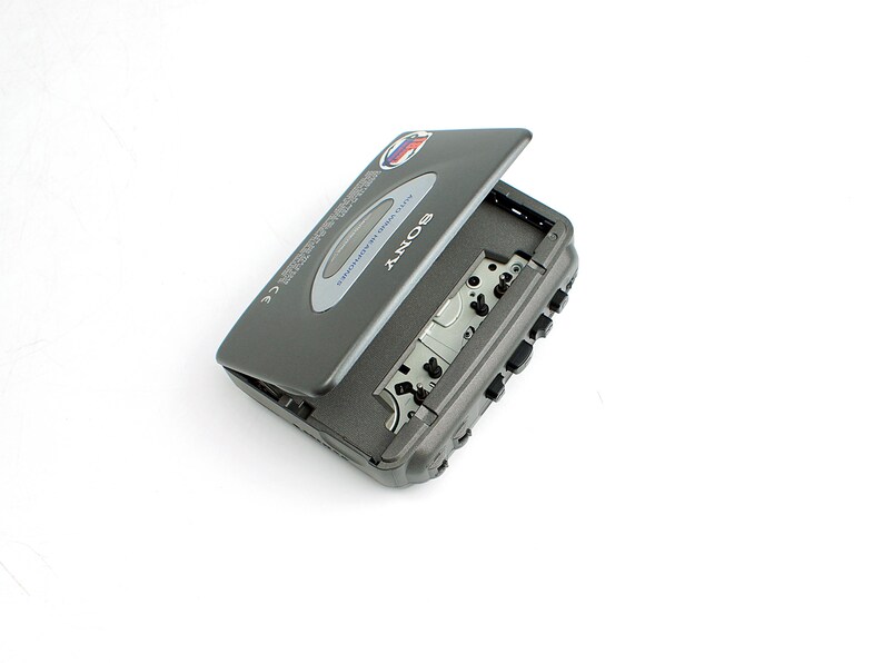 May include: A dark gray Sony Walkman cassette player with the cassette compartment door partially open. The device features a silver oval accent on the lid, black control buttons on the side, and internal mechanical components visible inside.