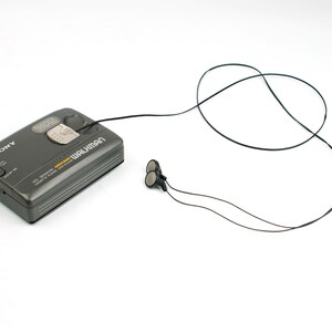 May include: A dark gray Sony Walkman cassette player with a rectangular body, featuring the brand name and model text printed on the top surface. A thin black cord connects the device to a pair of small, round black and silver wired earbuds.