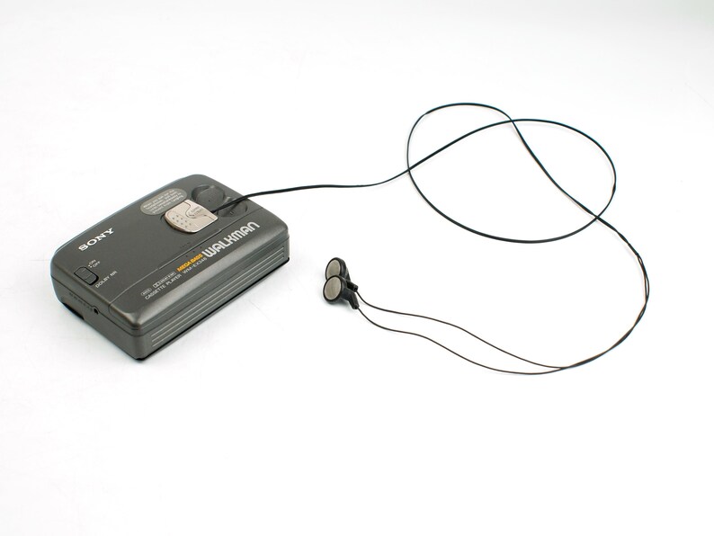 May include: A dark gray Sony Walkman cassette player with a rectangular body, featuring the brand name and model text printed on the top surface. A thin black cord connects the device to a pair of small, round black and silver wired earbuds.