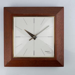 May include: Square wooden clock with a light-colored face and a dark brown wooden frame. The clock face has a minimalist design with thin black lines and the text "Dugena Atelier". The clock hands are black and silver.