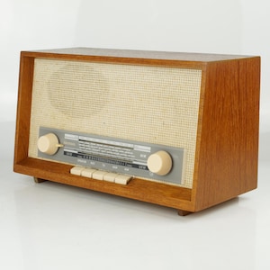 Puede incluir: Una radio de madera vintage con una cubierta de altavoz de color beige claro y un panel de control plateado. La radio tiene dos perillas de color crema y varios botones rectangulares. El marco de madera tiene un tono marrón rojizo cálido.