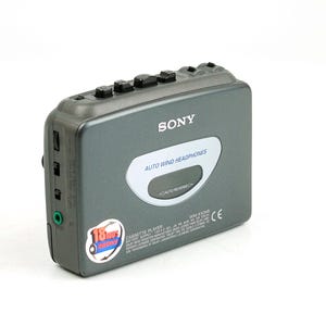 May include: A dark gray Sony cassette player featuring a top-mounted control panel with black buttons, a central oval display labeled Auto Wind Headphones, and a circular sticker on the lower left corner indicating 18 hours of battery life.