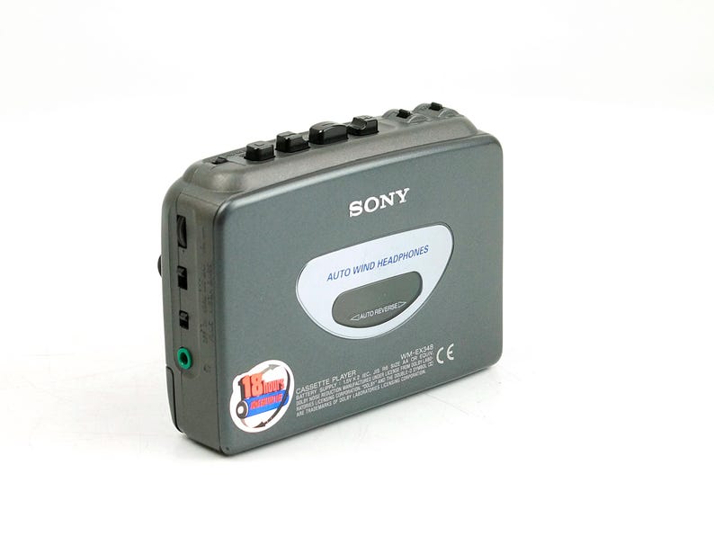 May include: A dark gray Sony cassette player featuring a top-mounted control panel with black buttons, a central oval display labeled Auto Wind Headphones, and a circular sticker on the lower left corner indicating 18 hours of battery life.