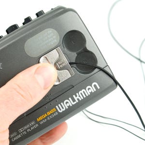 May include: A dark gray Sony Walkman cassette player model WM-EX348 with Mega Bass. The device features a silver Auto Wind mechanism, black control buttons, and a thin black cord extending from the side. Text on the device includes Sony, Walkman, Mega Bass, and Dolby B NR.