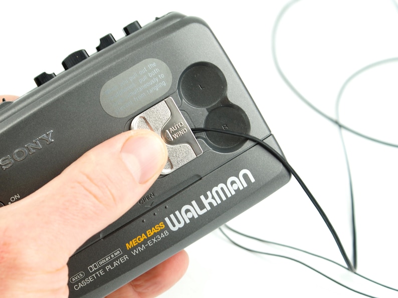 May include: A dark gray Sony Walkman cassette player model WM-EX348 with Mega Bass. The device features a silver Auto Wind mechanism, black control buttons, and a thin black cord extending from the side. Text on the device includes Sony, Walkman, Mega Bass, and Dolby B NR.