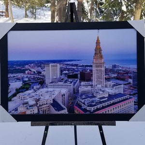 May include: A framed photograph of a cityscape with a tall building in the center. The building has a pointed roof and a clock tower. The city is bathed in a soft, purple light. The photograph is displayed on an easel.