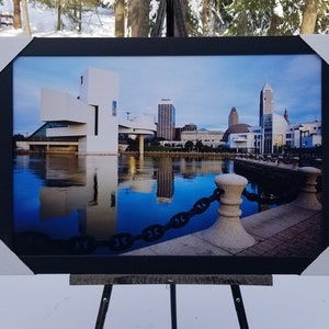 May include: A framed photograph of a cityscape with a modern building in the foreground. The building is white and has a unique design. The cityscape is reflected in the water in the foreground. The photo is framed in black and is on an easel.