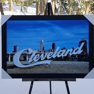 May include: A framed print of the Cleveland skyline with the city's name in white script. The image is displayed on a black easel against a snowy backdrop with trees. The sky is blue.
