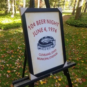 May include: A black framed sign with white text that reads "10&cent; Beer Night June 4, 1974 Cleveland, Ohio Municipal Stadium". The sign also features a black and white illustration of a baseball stadium.