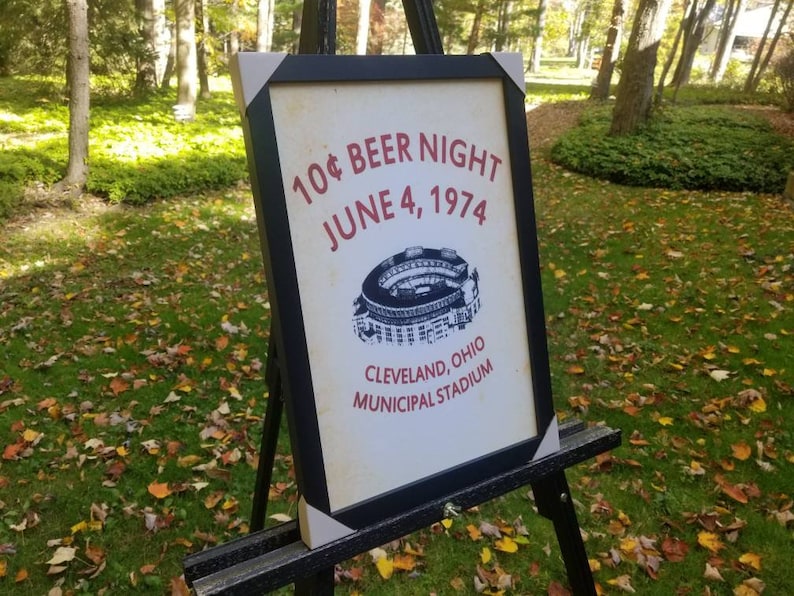 May include: A black framed sign with white text that reads "10&cent; Beer Night June 4, 1974 Cleveland, Ohio Municipal Stadium". The sign also features a black and white illustration of a baseball stadium.