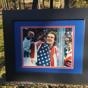 May include: Framed photograph of a smiling person draped in an American flag. The photograph is set within a blue and red mat, enclosed by a black frame. The background shows a crowd of people.
