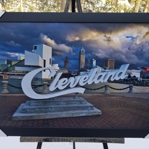 May include: Framed photograph of the Cleveland skyline with a large white sign that reads "Cleveland" in a stylized font. The sign is in front of the city's waterfront and the skyline is visible in the background. The sky is cloudy and the sun is setting.