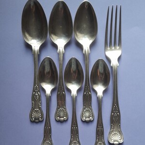 Op de afbeelding: Een set van zeven zilverkleurige bestekdelen, waaronder zes lepels en een vork. De lepels zijn van verschillende groottes, met sierlijke handvatten en een gepolijste afwerking. De vork heeft een klassiek ontwerp met vier tanden.