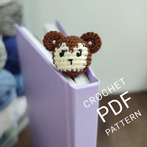 Może przedstawiać: Szydełkowy zakładka do książki w kształcie małpy w kolorach brązowym i kremowym, umieszczona na fioletowej książce. Zakładka ma szczegółową twarz z dużymi uszami. Tekst "CROCHET PDF PATTERN" jest widoczny na zdjęciu.
