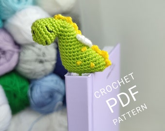 May include: A green crocheted dinosaur bookmark with yellow accents. The bookmark is sitting on a purple book with the text "CROCHET PDF PATTERN" printed on the cover.