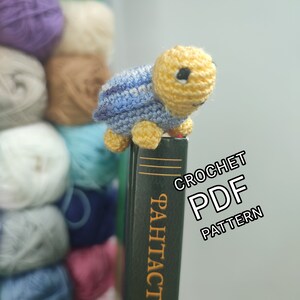 May include: A small, crocheted turtle with a yellow head and feet, and a blue and white striped shell. The turtle is perched on a dark green book. The text "CROCHET PDF PATTERN" is visible in the image. Yarn balls are in the background.