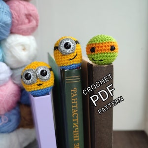 May include: Two crocheted character bookmarks, one yellow and one green, are placed on top of a stack of books. The yellow bookmark is a Minion character with a blue and yellow outfit and the green bookmark is a Teenage Mutant Ninja Turtles character with an orange mask. The books have the text "CROCHET PDF PATTERN" on the spine.