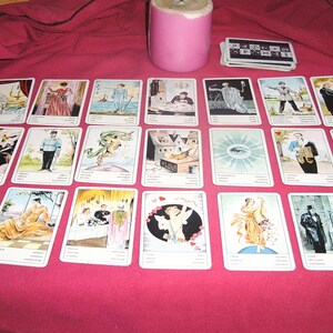 Pu&ograve; includere: Un insieme di carte dei tarocchi illustrate, disposte su un tessuto rosso. Sono presenti anche una candela rosa e un mazzo di carte. Le carte presentano varie figure e scene, suggerendo un tema di divinazione.