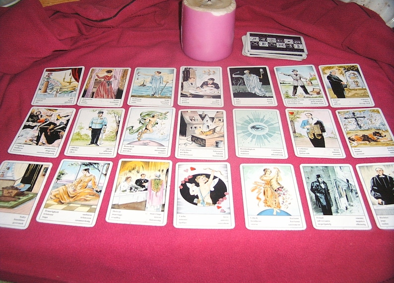 Pu&ograve; includere: Un insieme di carte dei tarocchi illustrate, disposte su un tessuto rosso. Sono presenti anche una candela rosa e un mazzo di carte. Le carte presentano varie figure e scene, suggerendo un tema di divinazione.