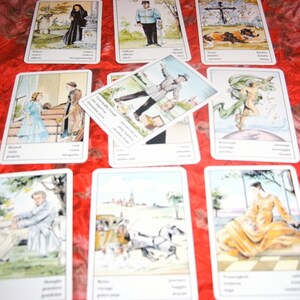 Pu&ograve; includere: Un insieme di carte dei tarocchi con illustrazioni e testo. Le carte presentano varie scene, tra cui figure in abiti storici, paesaggi e immagini simboliche. Le carte sono disposte su una superficie rossa a motivi.