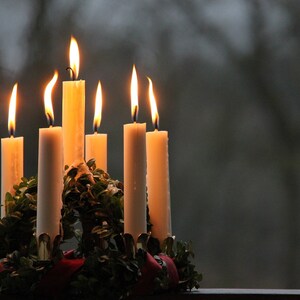 May include: A lit Advent wreath with seven white candles. The candles are arranged in a circle, with flames flickering against a blurred, dark background. The wreath is decorated with green foliage and a red ribbon.