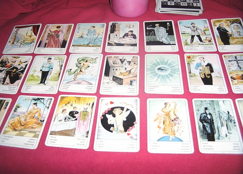 Pu&ograve; includere: Un insieme di carte dei tarocchi disposte su una superficie rosa. Ogni carta presenta un'illustrazione unica con testo. Le carte raffigurano varie scene e figure, con un'estetica vintage. Un contenitore rosa e un mazzo di carte sono visibili sullo sfondo.
