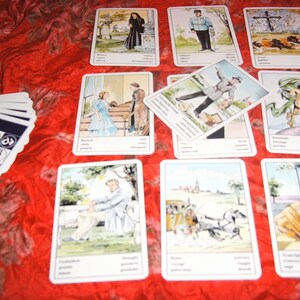 Pu&ograve; includere: Un mazzo di carte dei tarocchi con illustrazioni di persone e scene, disposte su una superficie rossa e strutturata. Le carte presentano immagini con testo in basso. &Egrave; visibile un mazzo di carte separato con simboli.