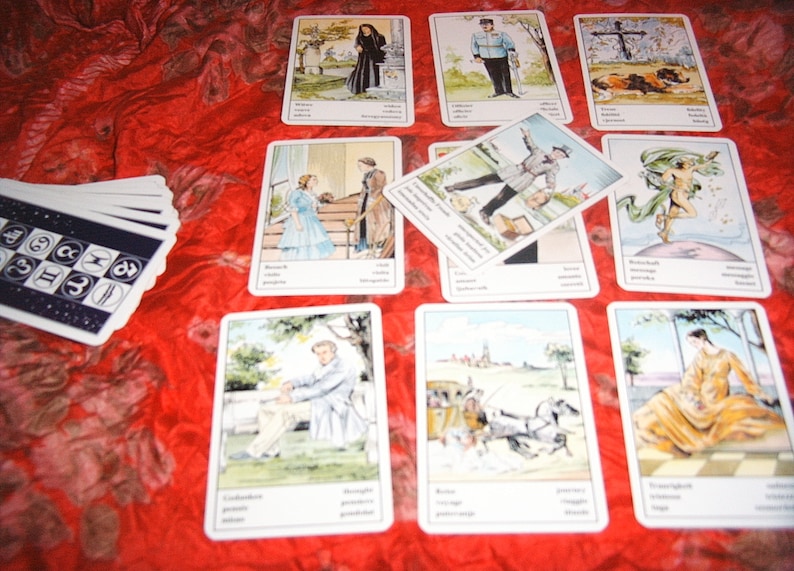 Pu&ograve; includere: Un mazzo di carte dei tarocchi con illustrazioni di persone e scene, disposte su una superficie rossa e strutturata. Le carte presentano immagini con testo in basso. &Egrave; visibile un mazzo di carte separato con simboli.