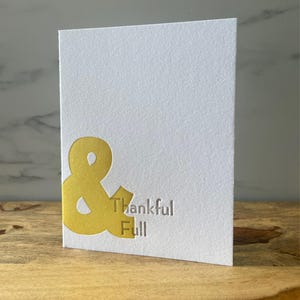 May include: A white card with a gold ampersand symbol and the text "Thankful & Full" printed in gray.