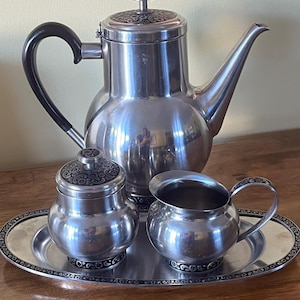 May include: A silver tea set comprising a coffee pot, creamer, sugar bowl, and serving tray. The coffee pot features a black handle and a decorative lid. The tray and lids have a black and silver ornate design. The set is on a wooden surface.