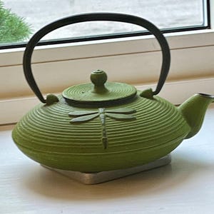 May include: A lime green cast iron teapot featuring a textured, ribbed surface and a raised dragonfly motif on the front. The teapot includes a matching circular lid with a small knob handle and a black, arched metal handle attached to the top.