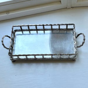 Peut inclure: Un plateau rectangulaire en métal argenté doté d'une base plate réfléchissante et d'une galerie surélevée au motif texturé rappelant le bambou. Le plateau comprend deux poignées incurvées assorties sur les côtés et repose sur de petits pieds.