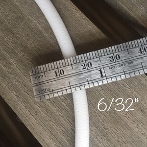 May include: A white, cylindrical foam tube is measured with a metal ruler. The ruler shows the tube is 6/32 inches in diameter. The tube is positioned on a wooden surface.