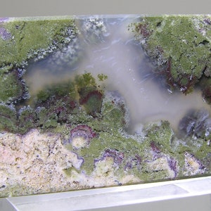 May include: A polished slab of agate with a unique pattern of green, white, and purple. The agate has a cloudy, translucent appearance with a variety of textures.