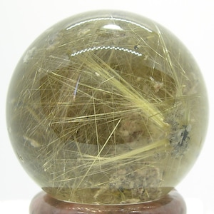 May include: A clear quartz sphere with golden rutilated inclusions. The sphere is approximately 2 inches in diameter and is displayed on a wooden base.