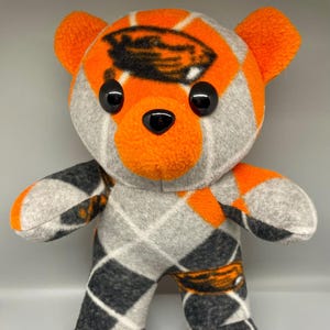 May include: A plush teddy bear with a gray argyle pattern, orange accents, and black eyes. The bear's head features an orange and black design. A cute, handmade stuffed animal.