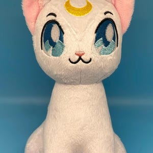 May include: A white plush cat with pink inner ears and blue eyes. A gold crescent moon is embroidered on its forehead. The cat is sitting upright on a blue background, with its paws tucked in front.