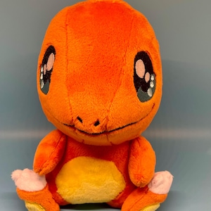 May include: An orange plush toy of a Charmander character with large, expressive eyes. The plush has a yellow belly and white accents on its feet. The toy is made of soft, fuzzy material and is sitting upright.