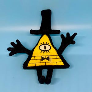 May include: A yellow triangle-shaped plush toy with a black top hat, black arms, and a single eye. The toy has a black bow tie and a brick pattern on its body.