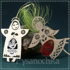 May include: Two wooden angel ornaments with intricate cutouts. One angel is holding a red egg with a bird design, and the other is holding a white egg with a bird design. The angels are both wearing a halo and have a smiling face.