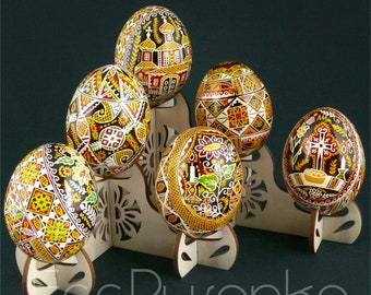 Könnte beinhalten: Fünf aufwendig bemalte ukrainische Pysanky-Eier, jedes mit einem einzigartigen Design, werden auf einem Holzständer präsentiert. Die Eier sind mit traditionellen Motiven in Rot, Gelb, Schwarz und Weiß verziert.