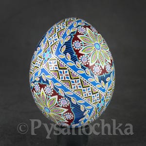 Op de afbeelding: Een handbeschilderd decoratief ei met ingewikkelde bloemen- en geometrische patronen. Het ei heeft een levendig kleurenpalet van blauw, bordeaux, geel en wit. Het ontwerp omvat gestileerde bloemen, geometrische vormen en bladmotieven.
