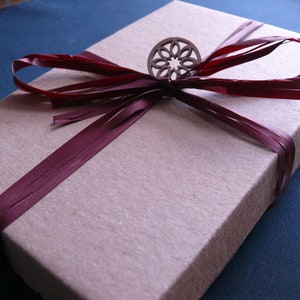 May include: A beige rectangular gift box tied with a deep burgundy ribbon. A decorative wooden embellishment is centered on the ribbon. The box is on a dark blue surface, suggesting a presentation or display.
